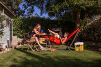 Girl lounging in Swiftlet portable hammock stand with Ember Oranger Roo Double hammock in the backyard playing games with friend 