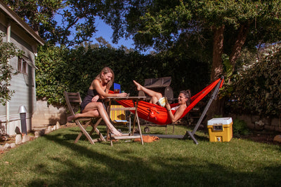Girl lounging in Swiftlet portable hammock stand with Ember Oranger Roo Double hammock in the backyard playing games with friend