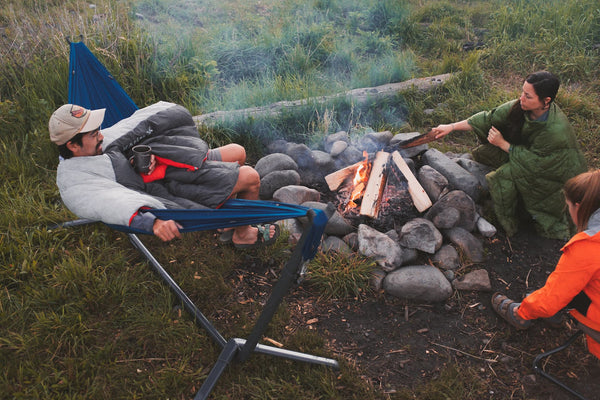 Three people sitting around a firepit made of stones. One man is relaxing in a hammock on a Kammok Swiftlet Portable Hammock Stand.