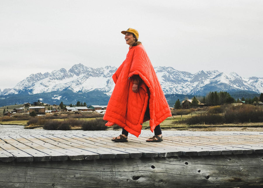 Woman walking with snowapped mountains and vilage in the distance wearing a Kammok Trail Quilt Firebelly 30°F in Poncho mode.