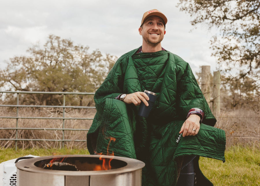 Man wearing a Kammok Mountain Blanket in poncho mode sitting in front of a fire outside.