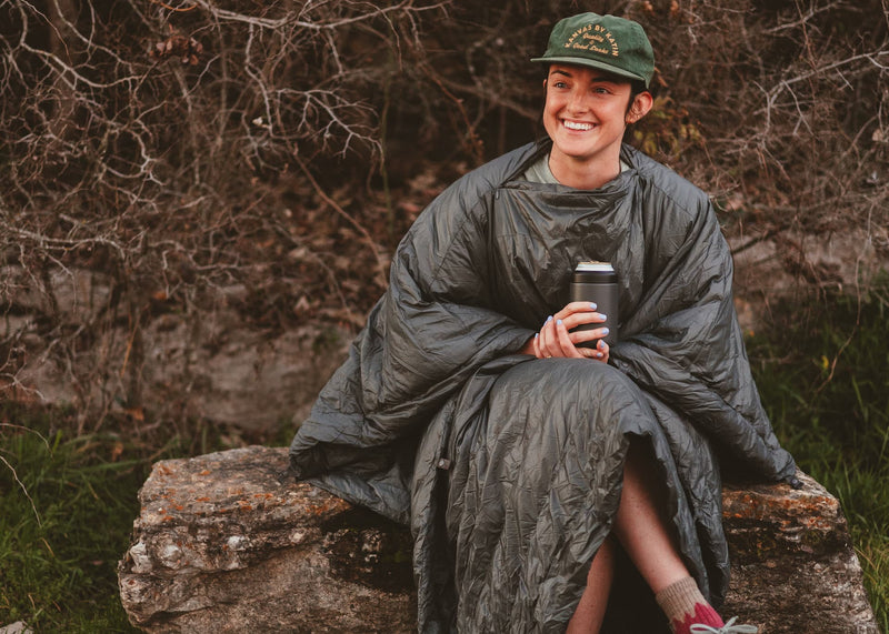 Girl sitting on a rock wearing a Kammok Trail Quilt Firebelly 30°F in Poncho Mode.