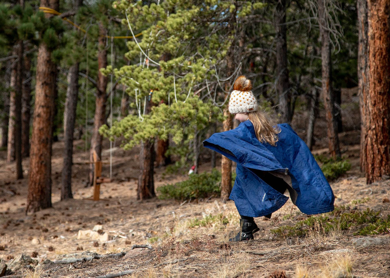 Little girl in a winter hat running through the woods wearing a Kammok Field Blanket Jr. in Poncho mode.