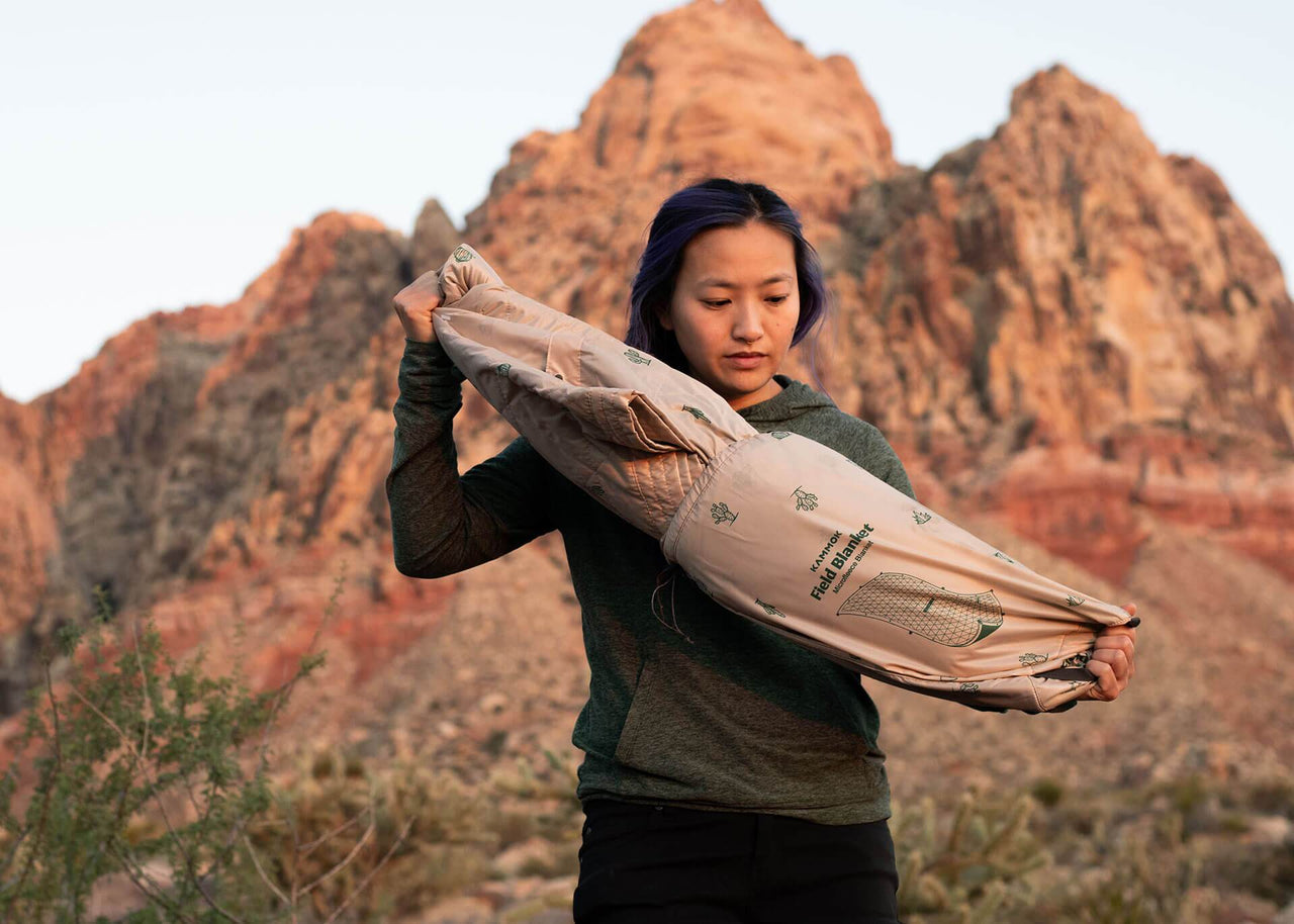 Woman pulls a Kammok Field Blanket Printed in mustard sand tan pink color with cacti print out from a water-resistant storage stuff sack near the desert mountains.