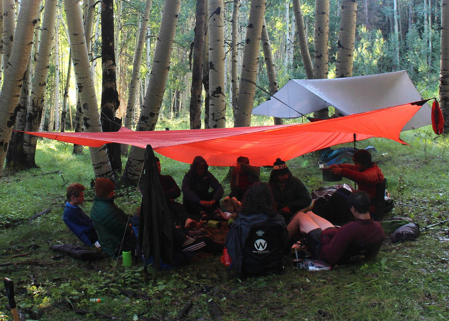 Large croup of people huddled underneath a Kammok Weather Shelter Kuhli XL in the middle of the woods.