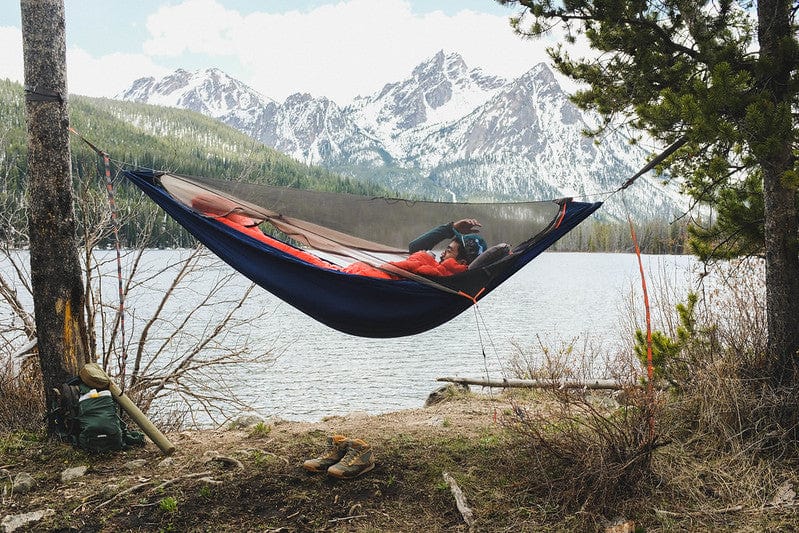 Man lying next to lake in a Kammok Hammock Tent Mantis with mountains in background.