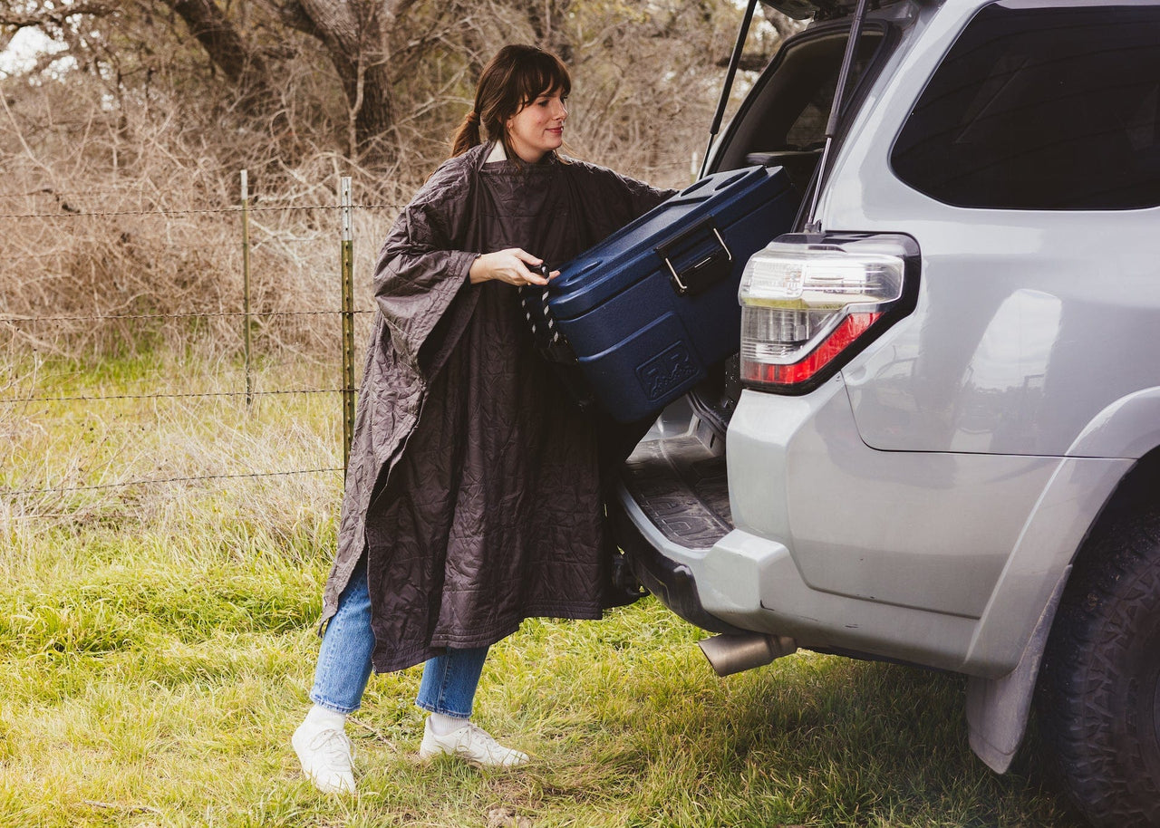 Women wearing a Kammok Field Blanket in poncho mode and loading her cooler into SUV.