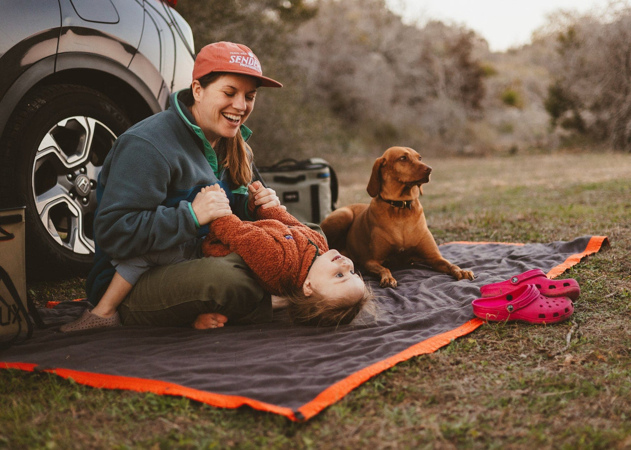 Mom sitting on Kammok Field Blanket with child leaning back out of her lap and dog lying next to them.