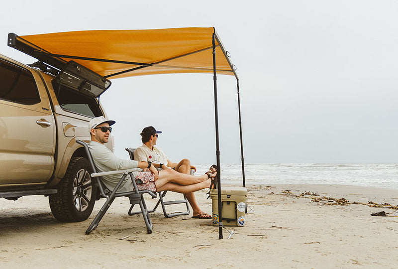 Two guys sitting on a beach in front of their truck underneath a Crosswing Shade Awning.