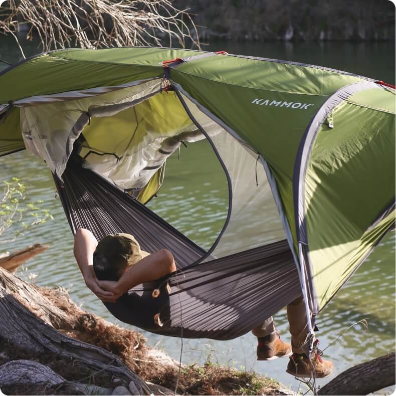Man overlooking the water while lounging in a Kammok Sunda Hammock Tent suspended between two trees.