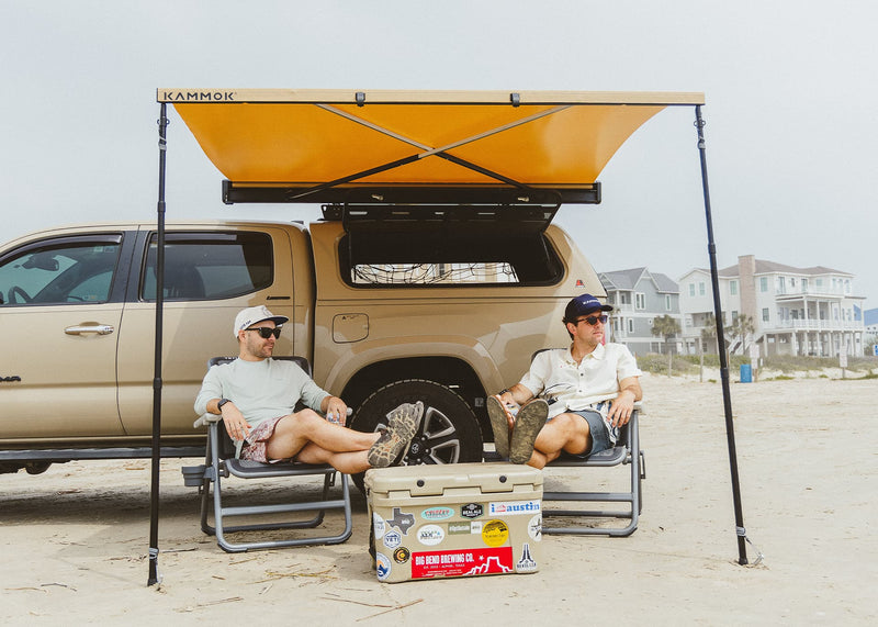 Two men on the beach sitting next to their truck underneath a Crosswing Awning with Pole Pack.