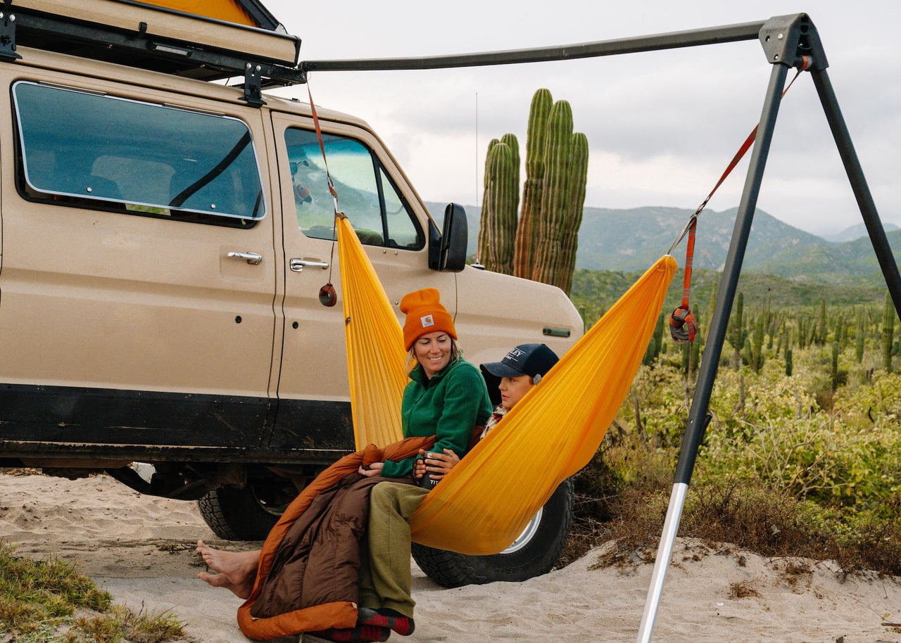 Mom & Child Sitting in a Roo Hammock on a Kammok Stand Outpost installed on a van with cacti and mountains in the background.