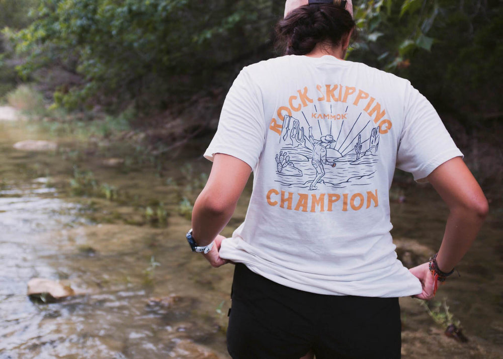 Women with back facing the camera wears a white shirt with the graphic Kammok Rock Skipping Champion. 