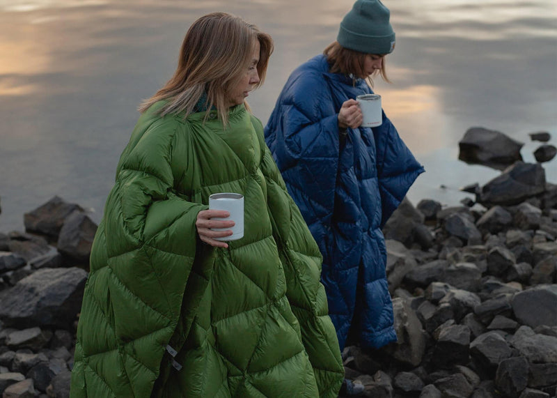 Two women walking on the rocks next to a lake with coffee waring their Bobcats in poncho mode.
