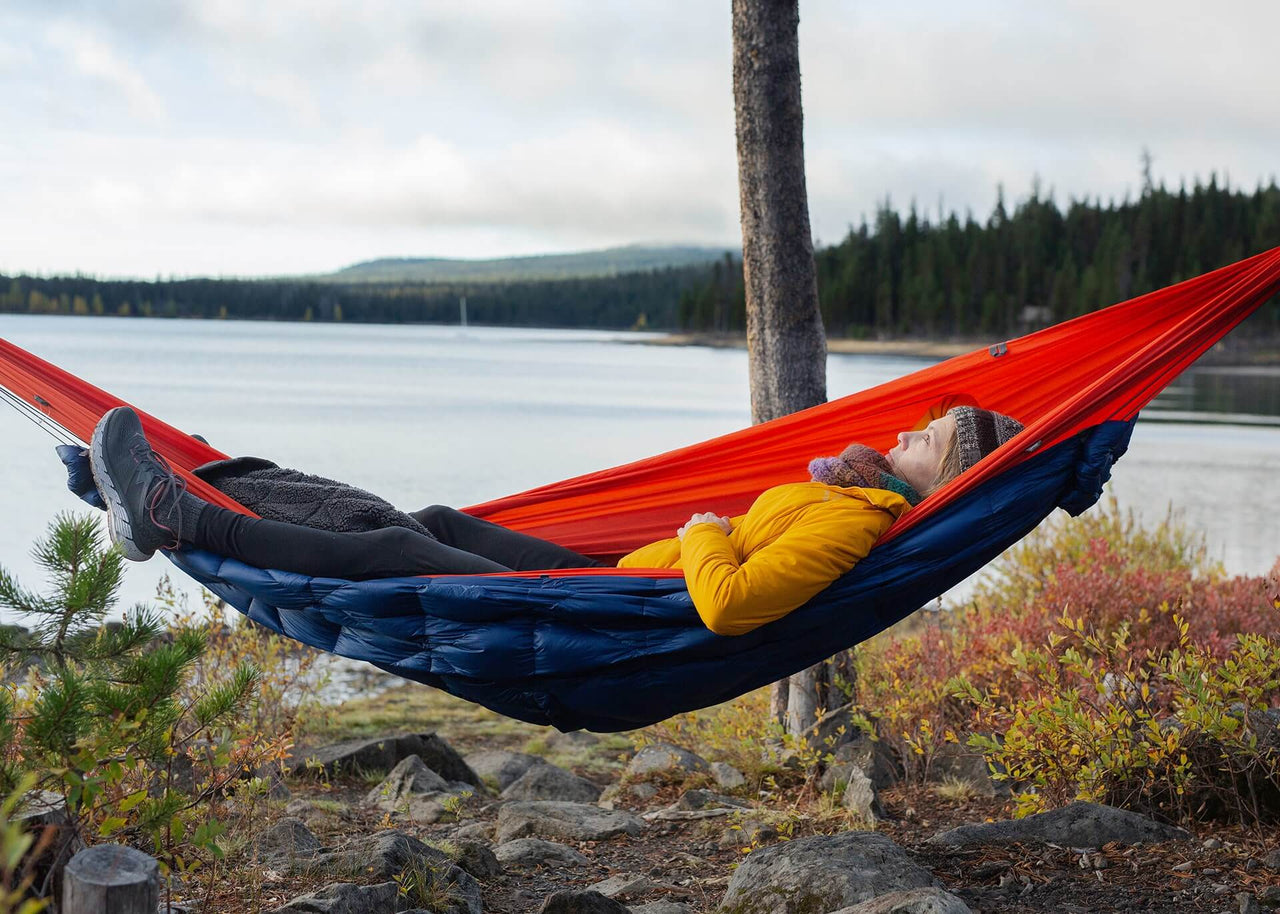 Woman laying in a hammock with her Kammok Trail Quilt Bobcat 45°F as an underquilt next to a lake.