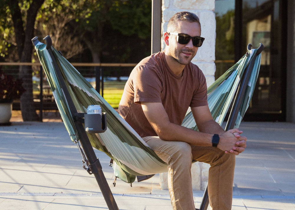 Man sitting in a Kammok Swiftet portable hammock stand in chair mode near the pool in a backyard with the Swiftlet cup holder attached.