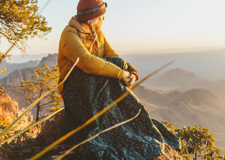 Women sitting in mountains with a winter hat overlooking the view wrapped in a Kammok Blanket Field Blanket Printed