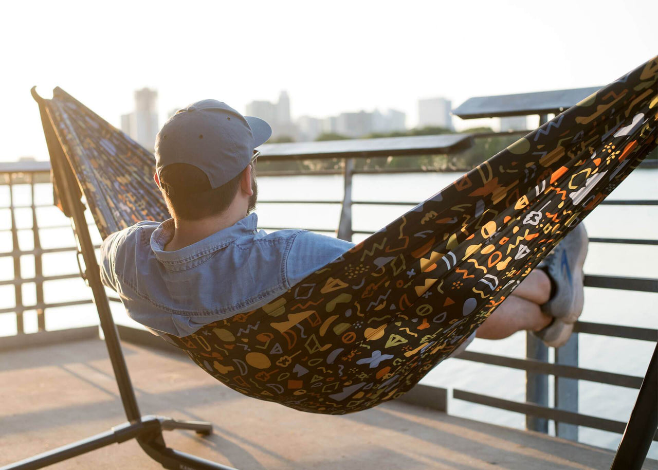 Man on back deck sitting on Kammok Hammock Roo Double Printed attached to a Swiftlet Hammock stand with skyline in distance.