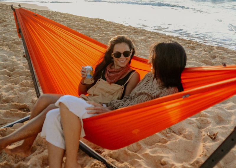 Two girls sitting in a Kammok Hammock Roo Double on a Swiflet Portable Hammock Stand on the beach.