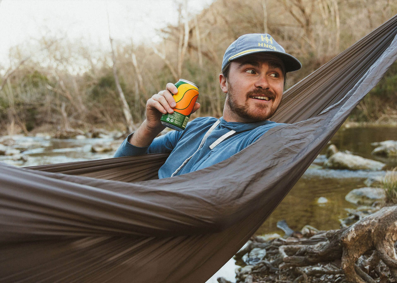 Man drinking a can while lying on a Kammok Hammock Roo Double XL Granite Gray next to the river.