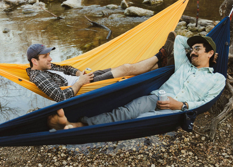 Two guys hanging side by side drinking beer at the river in their Kammok Hammock Roo Singles.